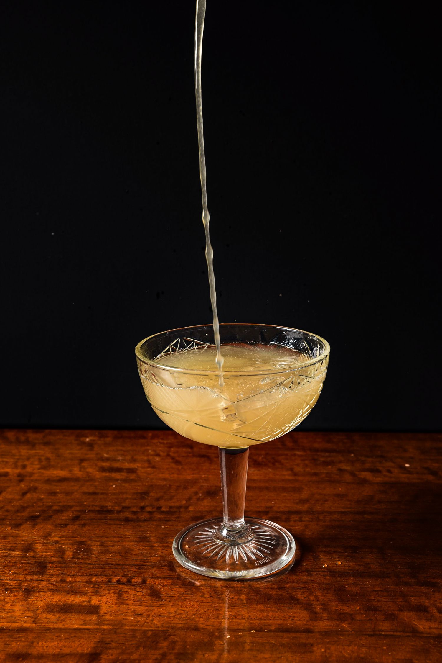 A sequence of four photos showing champagne being poured into a vintage coupe glass on a wooden table against a dark background.