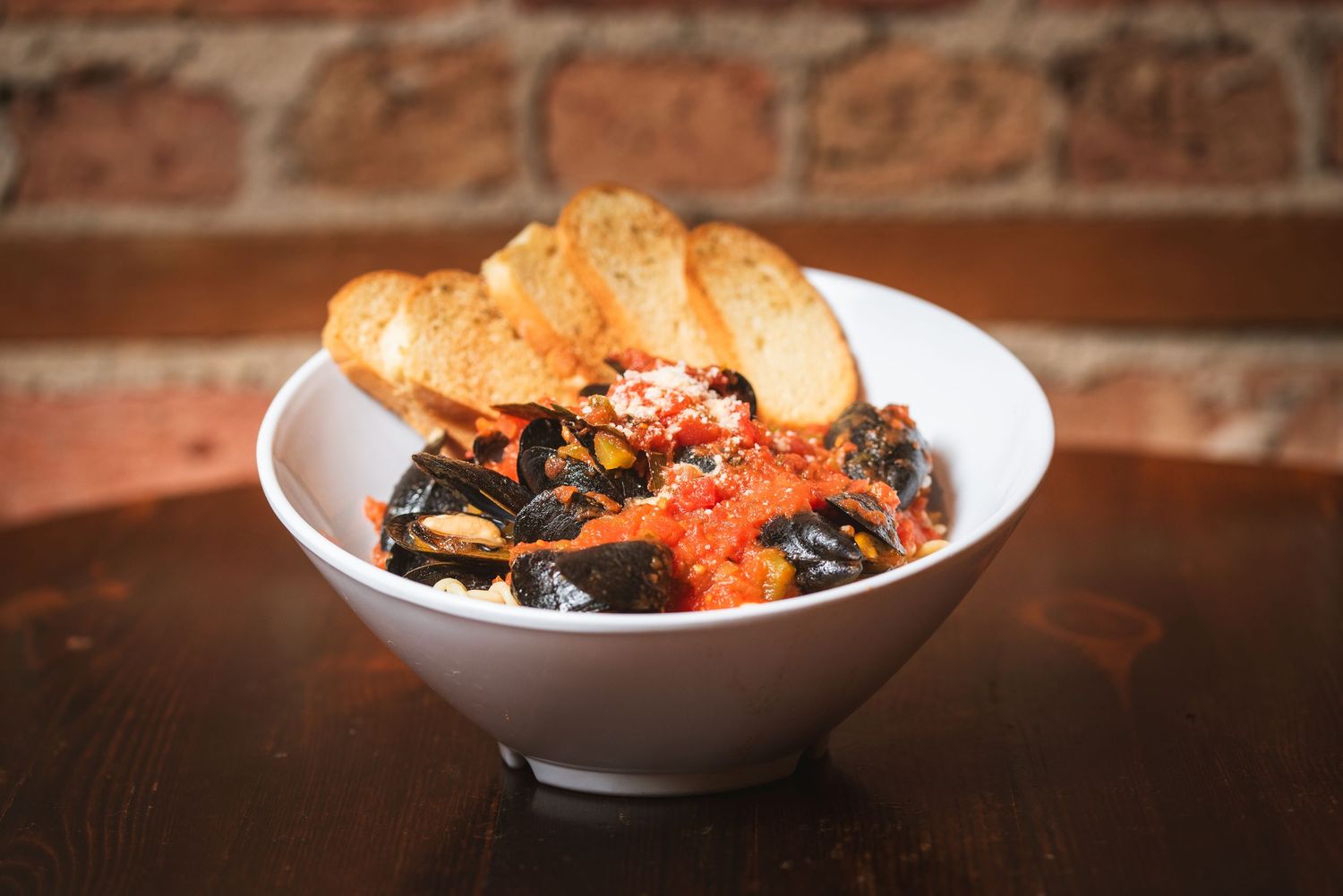 Bowl of steamed mussels in tomato sauce served with crusty bread on wooden table against brick wall background. From Embers in Lockport IL.
