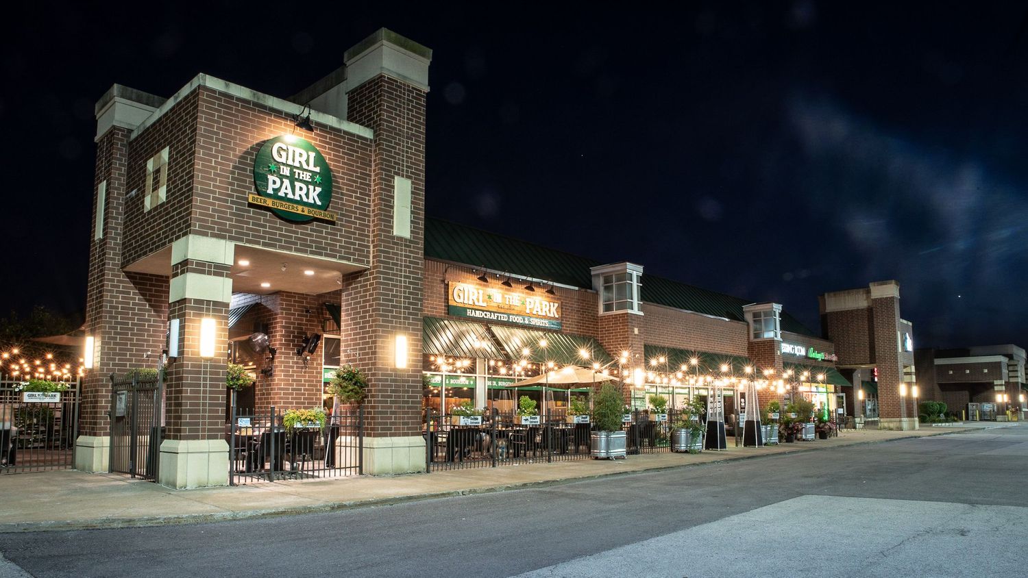 A panoramic nighttime view of a shopping center with illuminated storefronts and empty parking lot. Girl in The Park in Orland Park IL.