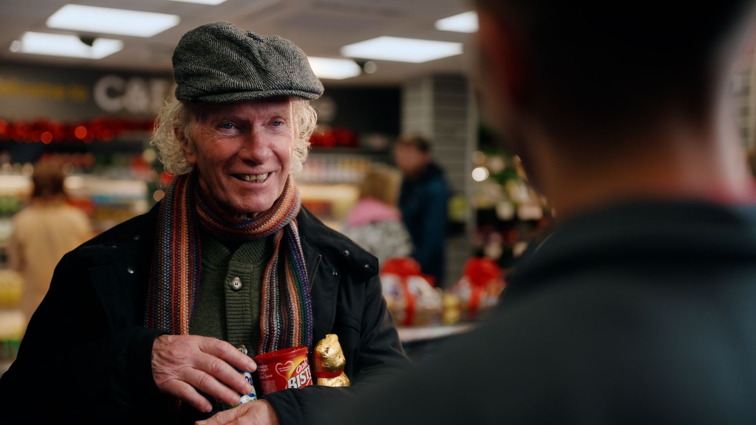 Smiling older man in flat cap and striped scarf holding items in a bright grocery store. Nearby Stores, S&W wholesale, TV Advert