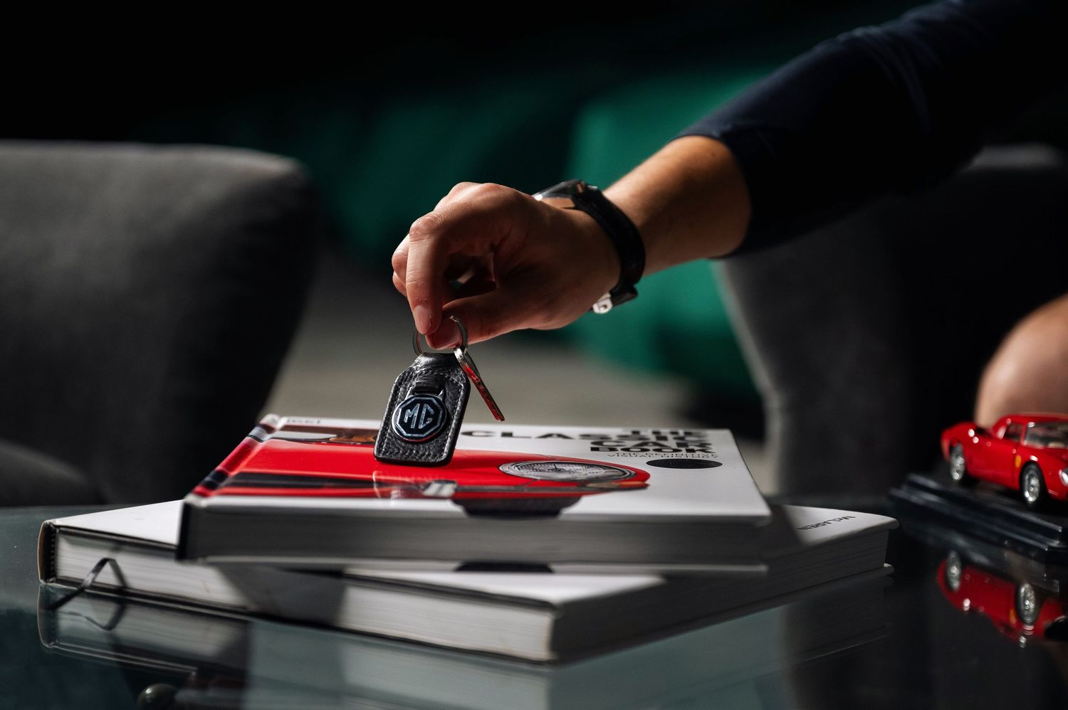 Hand placing a vintage MG car key on a red automotive book on a glass table. Motorist Collection