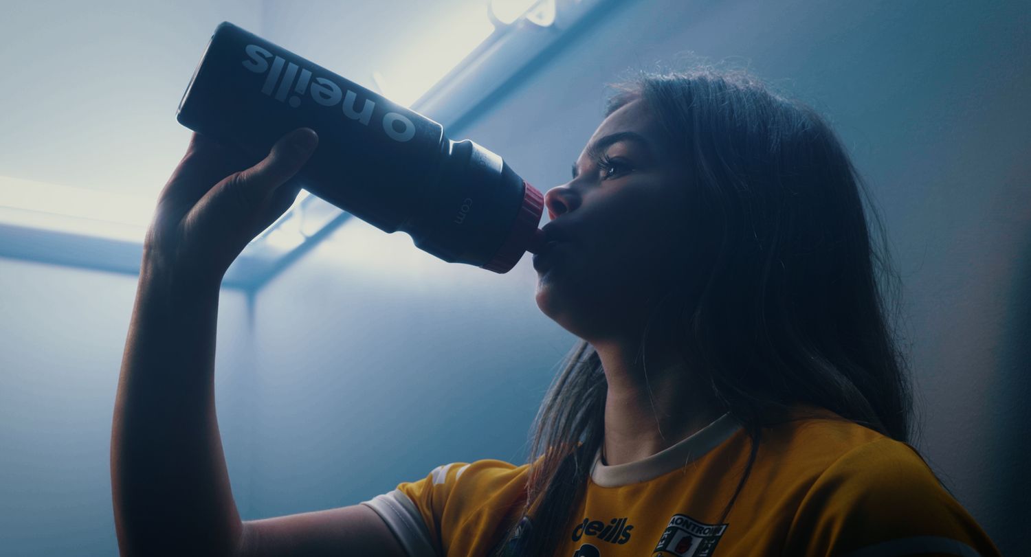 Young woman drinking from a black energy drink bottle under blue lighting. Ulster GAA