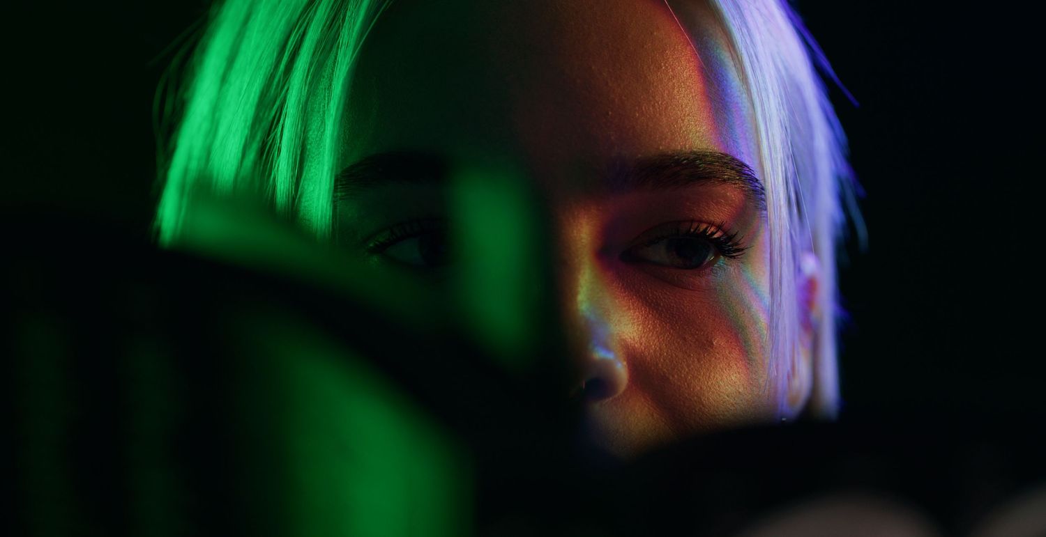 Close-up portrait of a woman with blonde hair illuminated by green and purple neon lights. Kaizen Brand & Print. Slims Kitchen