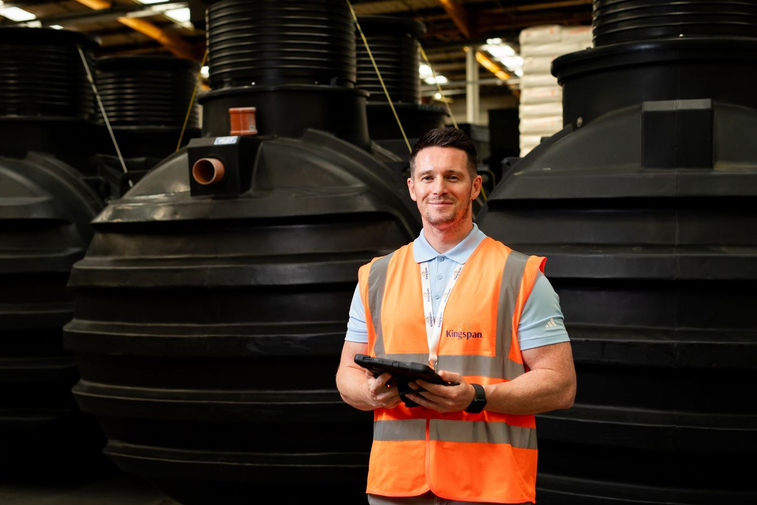 Kingspan warehouse employee in orange hi-vis vest holding tablet surrounded by large black storage tanks. Kingspan Water & Energy
