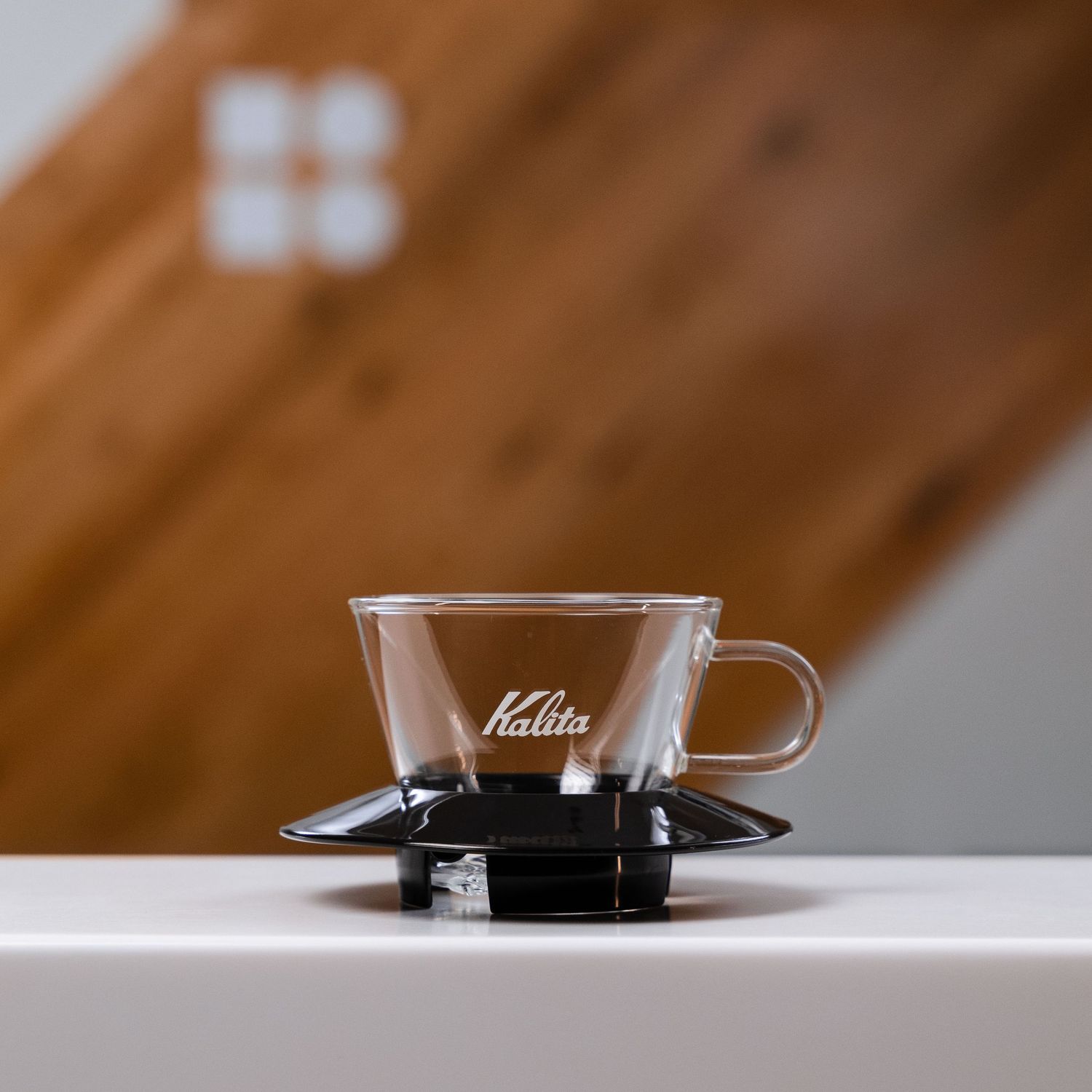 Kalita glass pour-over coffee dripper on a black stand displayed on a white surface. Bailies Coffee Roasters