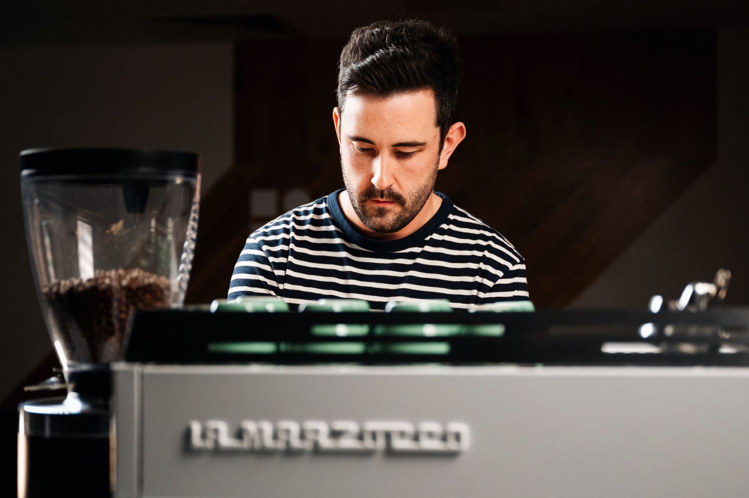 Male barista in striped shirt operating a La Marzocco espresso machine in a coffee shop. Bailies Coffee Roasters