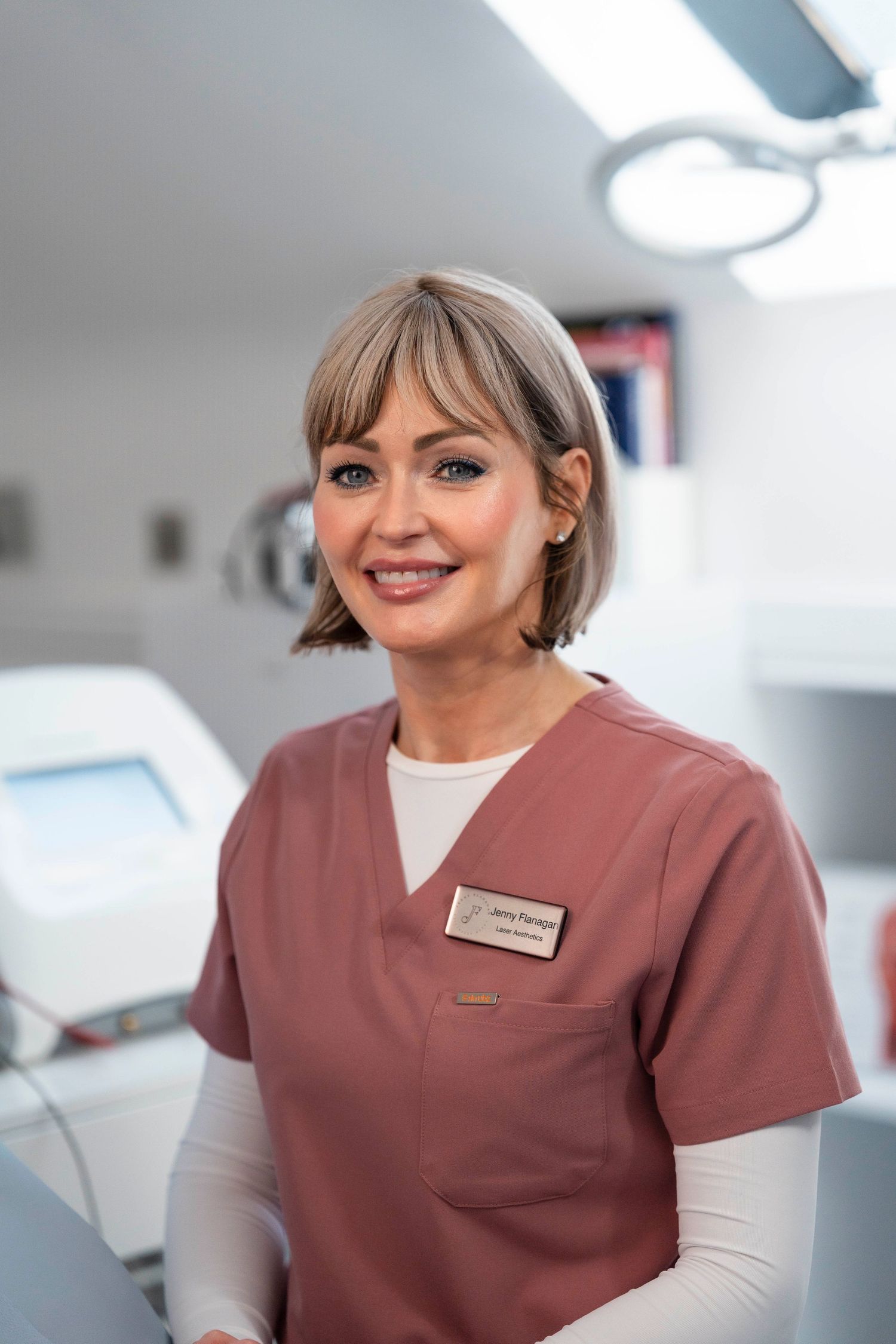 Smiling female healthcare professional in pink scrubs with name badge standing in modern medical clinic. Jenny Flanagan