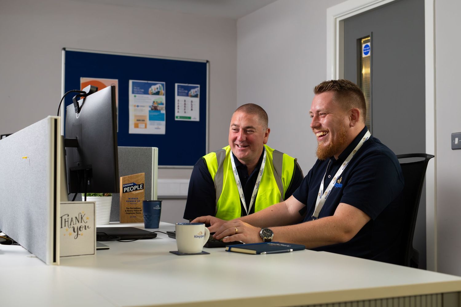 Two men smiling and laughing together at an office reception desk, one wearing a yellow high-visibility vest. Bailies Coffee Roasters