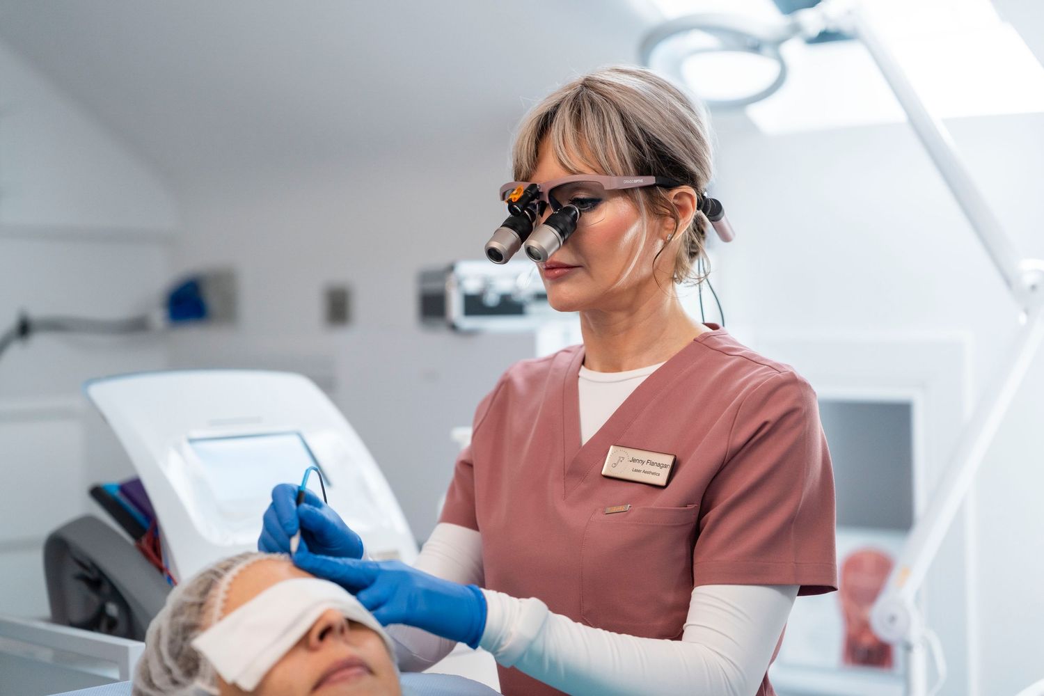 Jenny Flanagan aesthetic practitioner wearing magnifying loupes performs precision facial treatment on clinic patient.