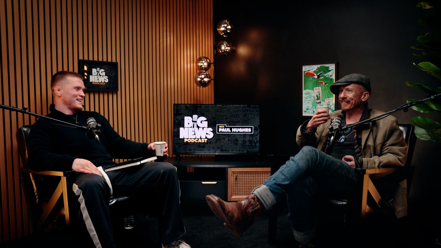 Two men sit in a dimly lit podcast studio engaged in conversation with a green book visible. Record Room, Big News Hughes Podcast
