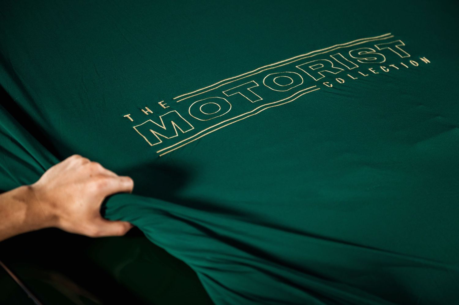 Hand pulling back a dark green cover revealing The Motorist Collection gold embroidered logo. Motorist Collection