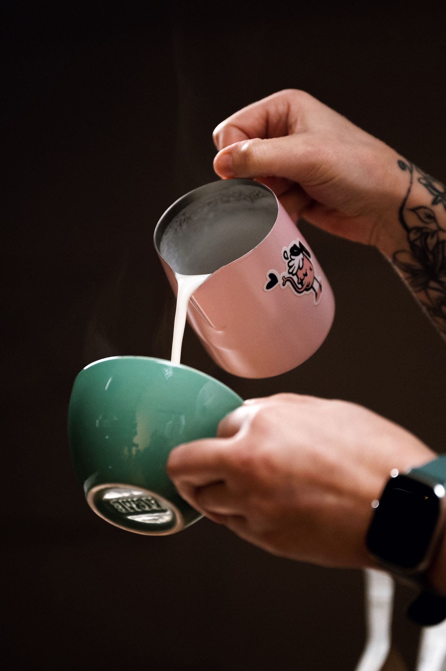 Tattooed barista pouring steamed milk from a pink pitcher into a teal coffee cup. Bailies Coffee Roasters