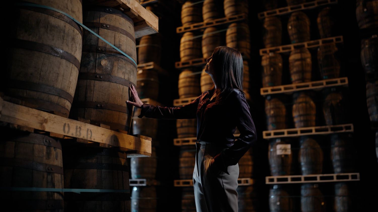 Person in hoodie inspecting wooden whisky barrels stacked on shelves in a dimly lit distillery warehouse. Bushmills Whiskey