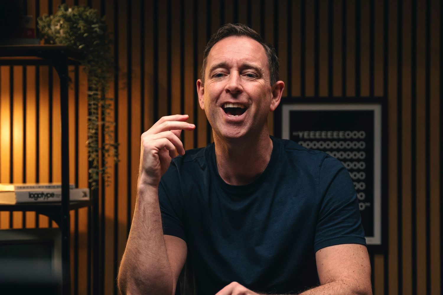 Connor McAuley in blue t-shirt laughing and gesturing in a modern studio with wooden panel walls.
