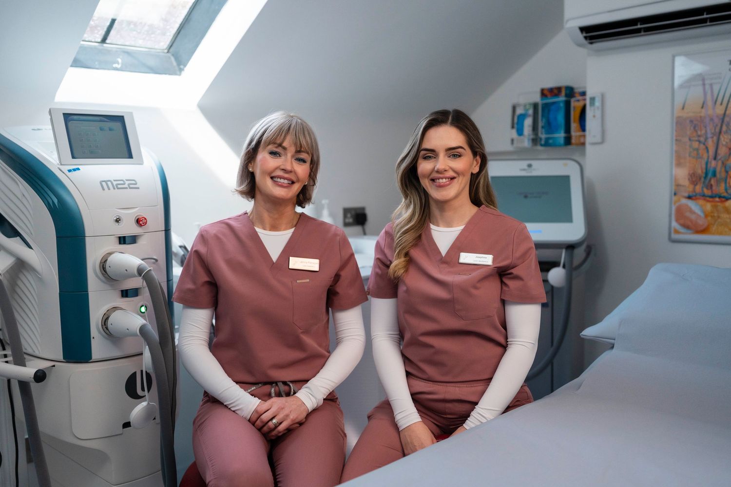 Two female medical professionals in pink scrubs smiling together in modern laser treatment room. Jenny Flanagan