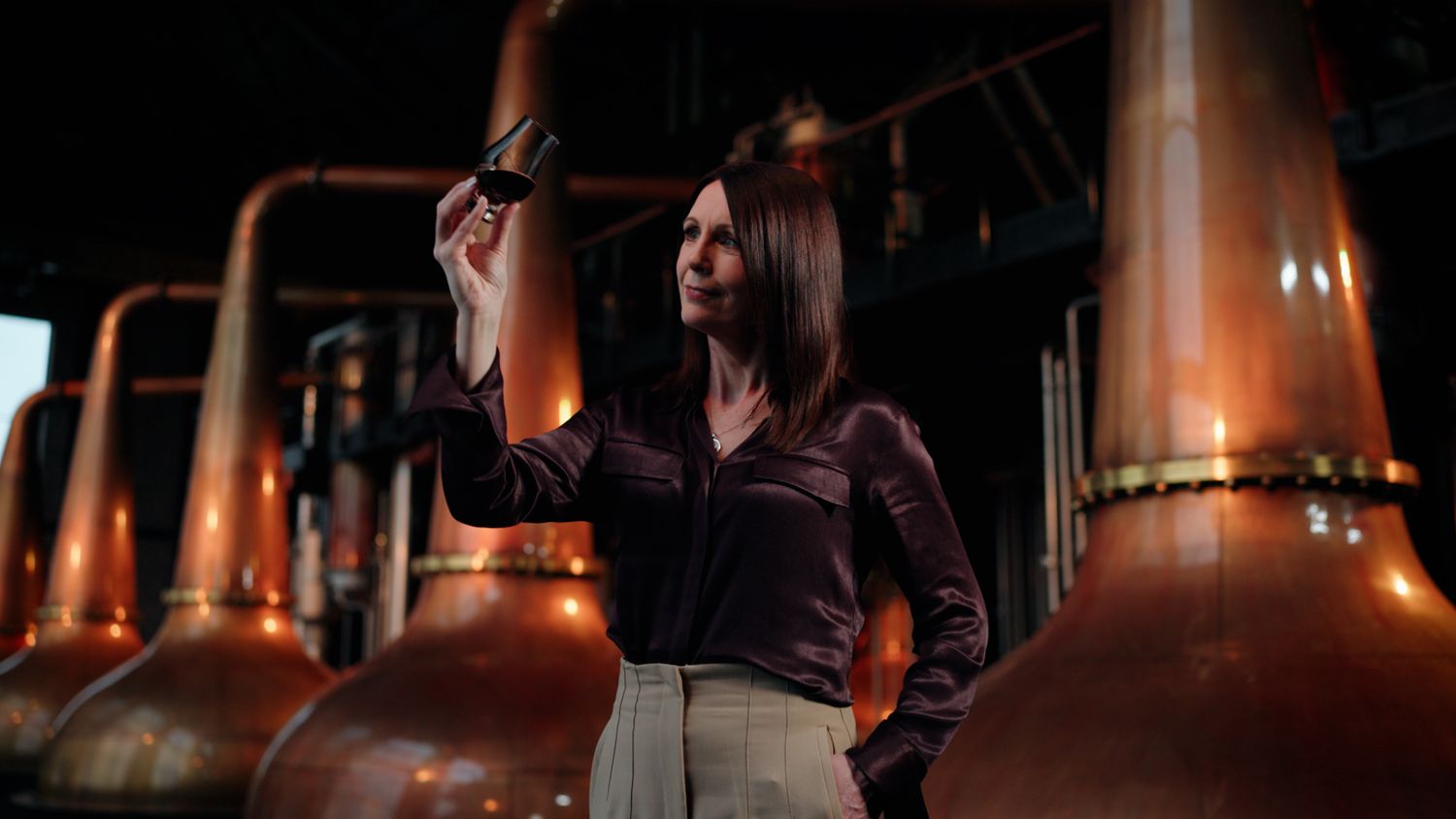 Woman examining whiskey sample beside gleaming copper stills in a distillery. Bushmills Whiskey