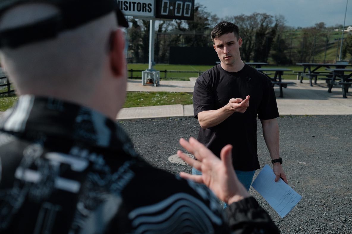 Two men discuss a film shoot outdoors near a sports scoreboard on a sunny day. Ross Reaney