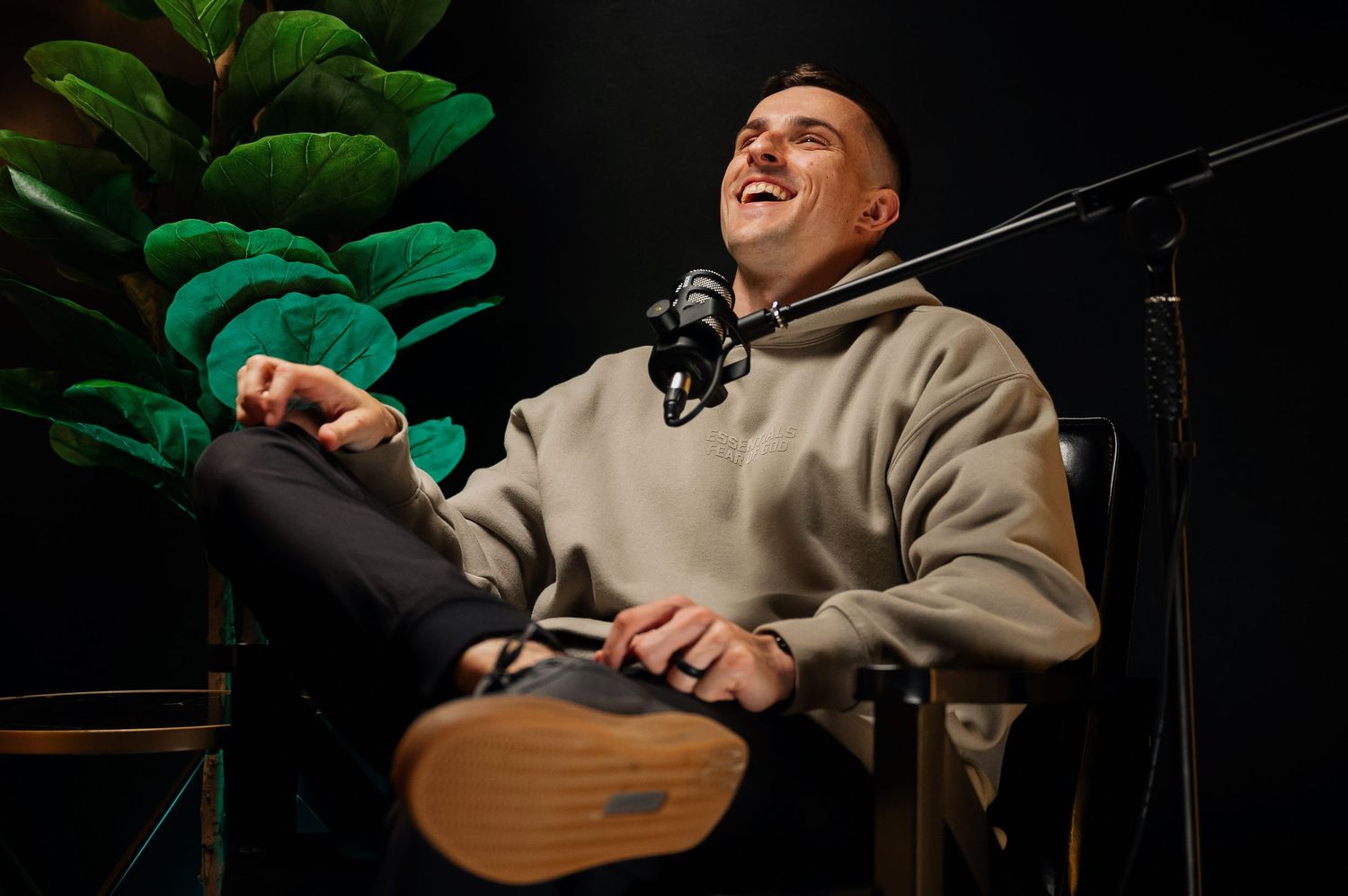 Man in beige hoodie laughing while sitting in podcast chair with microphone in dark studio.