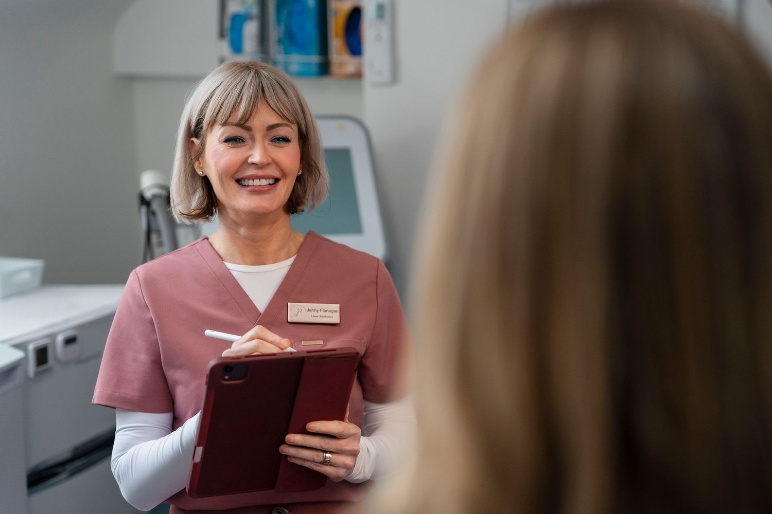 Jenny Flanagan aesthetic nurse in pink scrubs smiles while holding clipboard during patient consultation in clinic.