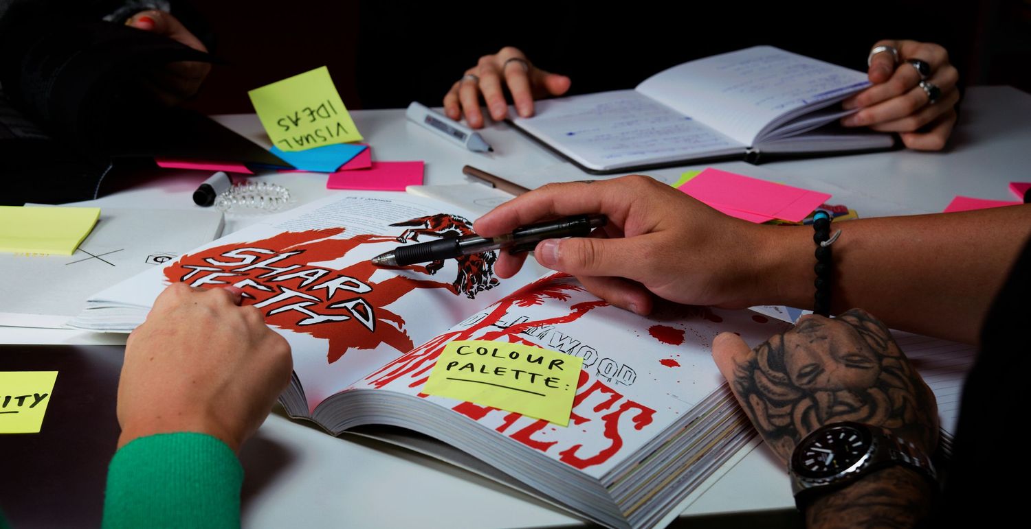Tattooed hands working on graphic design layout with colorful sticky notes and open books. Kaizen Brand & Print