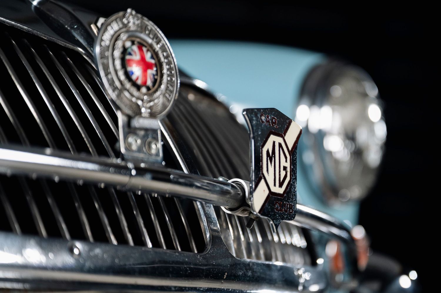 Close-up of chrome MG badge and grille on a classic light blue MGA sports car with club emblem. Motorist Collection
