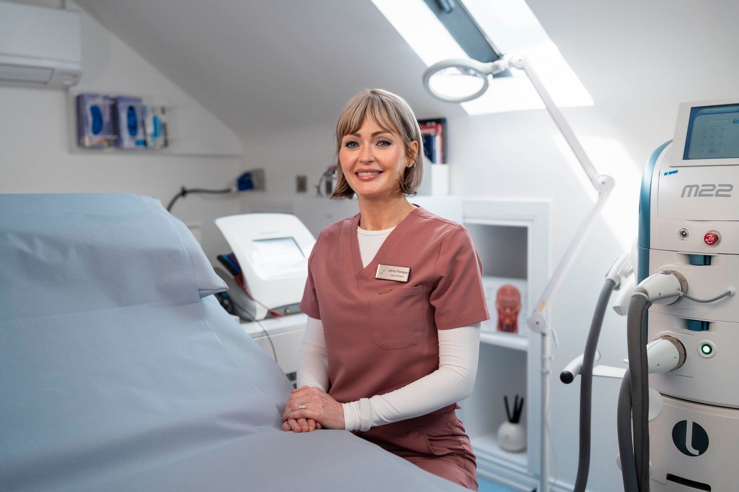 Blonde female aesthetician in pink scrubs posing beside a treatment bed in a modern clinic. Jenny Flanagan