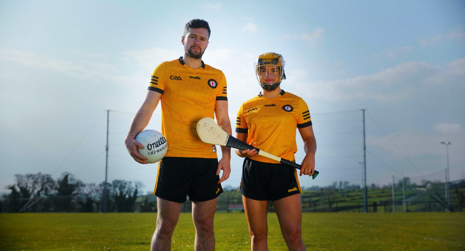 Two GAA players in yellow Antrim jerseys posing with a football and hurley on a pitch. Ulster GAA
