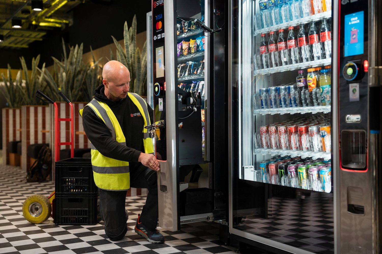 Worker in hi-vis vest restocking a modern vending machine with drinks and snacks in a commercial space.