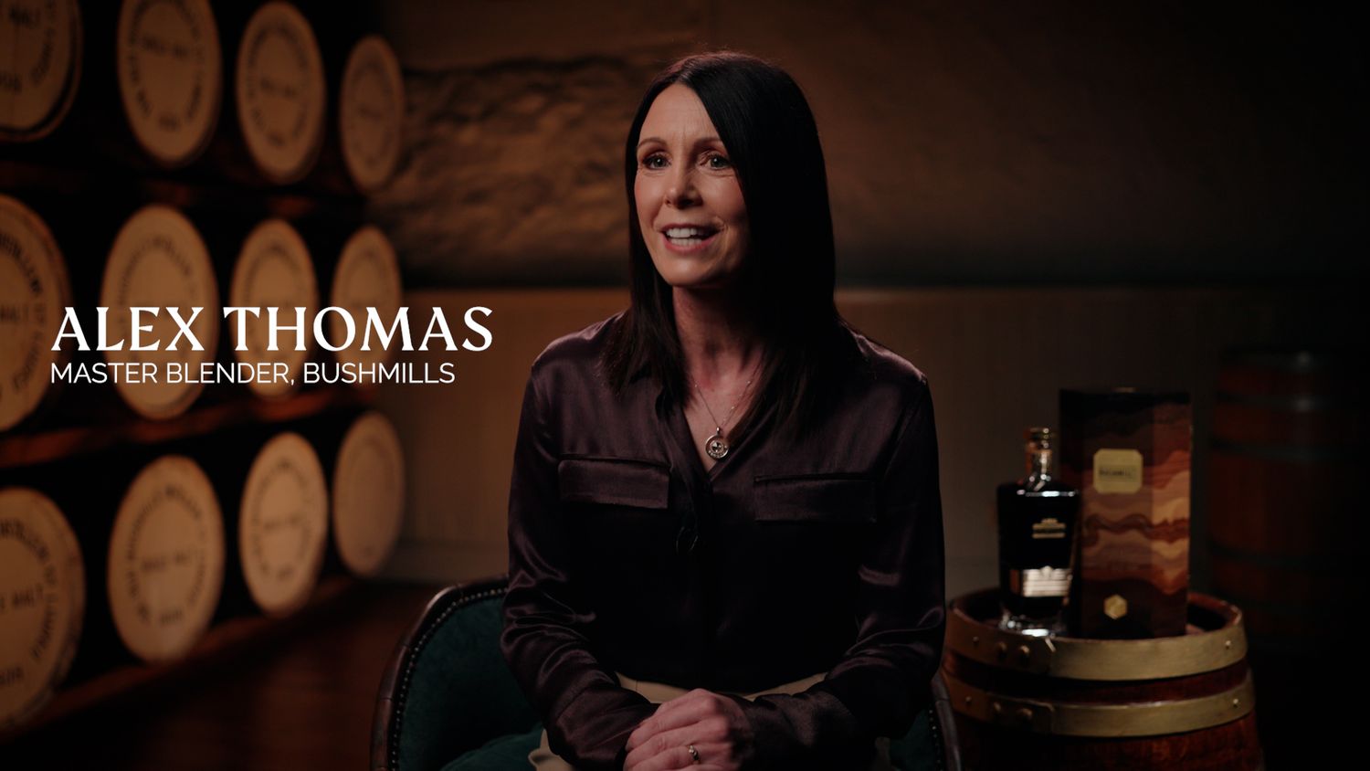 Alex Thomas, Master Blender at Bushmills, sits beside a whiskey barrel and bottle in a dimly lit distillery.  Bushmills Whiskey
