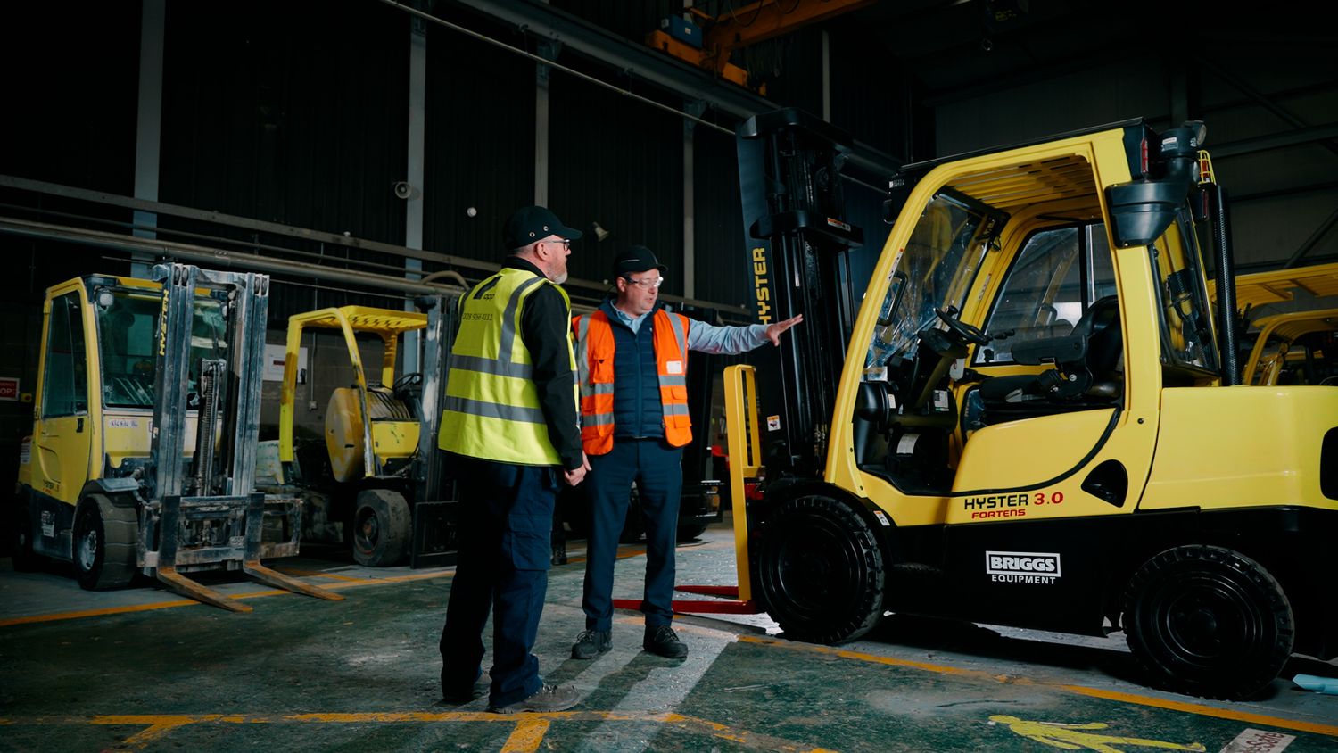 Two workers in hi-vis vests discussing a yellow Hyster forklift truck inside a warehouse facility. Briggs Equipment