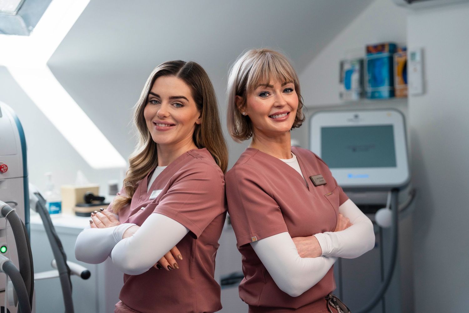 Two female medical aestheticians in pink scrubs pose back-to-back smiling in a modern clinic treatment room. Jenny Flanagan
