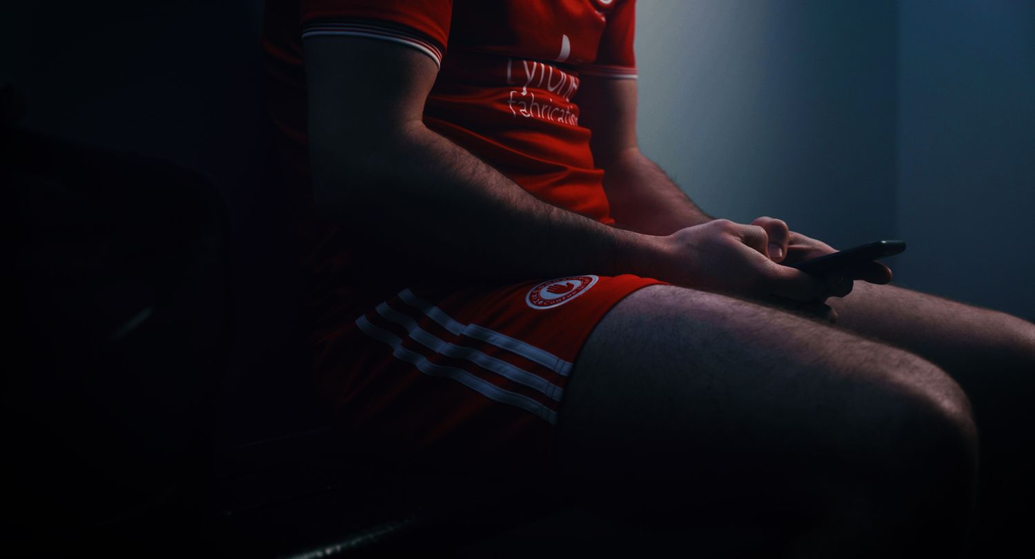Athlete in red Adidas GAA jersey and shorts holding a game controller in a dark room. Ulster GAA