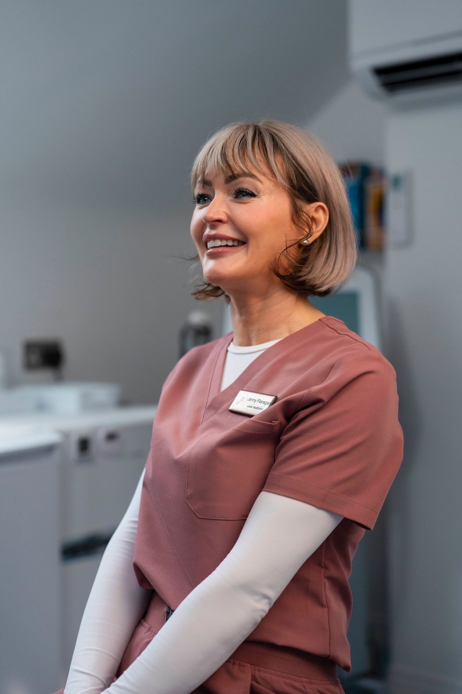Smiling Jenny Flanagan aesthetic practitioner in pink scrubs stands in modern medical clinic setting.