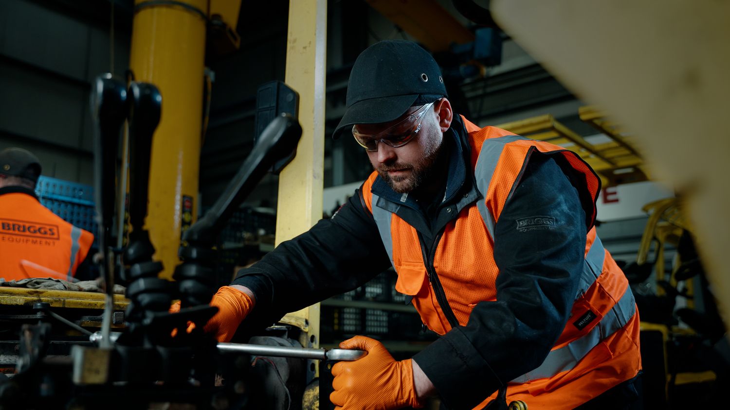 Industrial worker in orange hi-vis vest and black beanie performing maintenance on heavy machinery indoors. Briggs Equipment