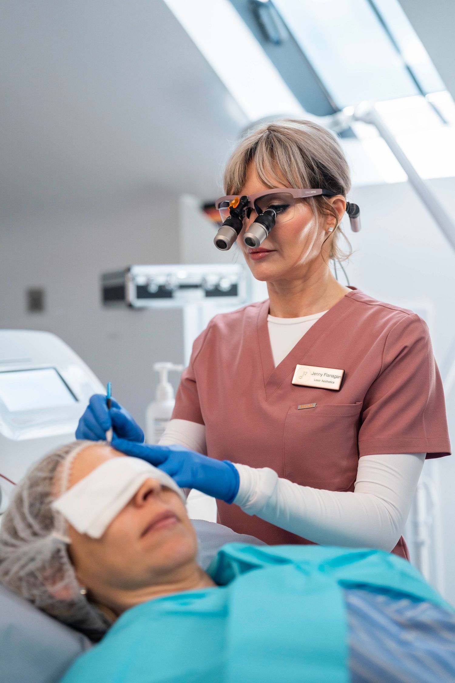 Jenny Flanagan doctor wearing surgical loupes performs facial treatment on patient in modern clinic.