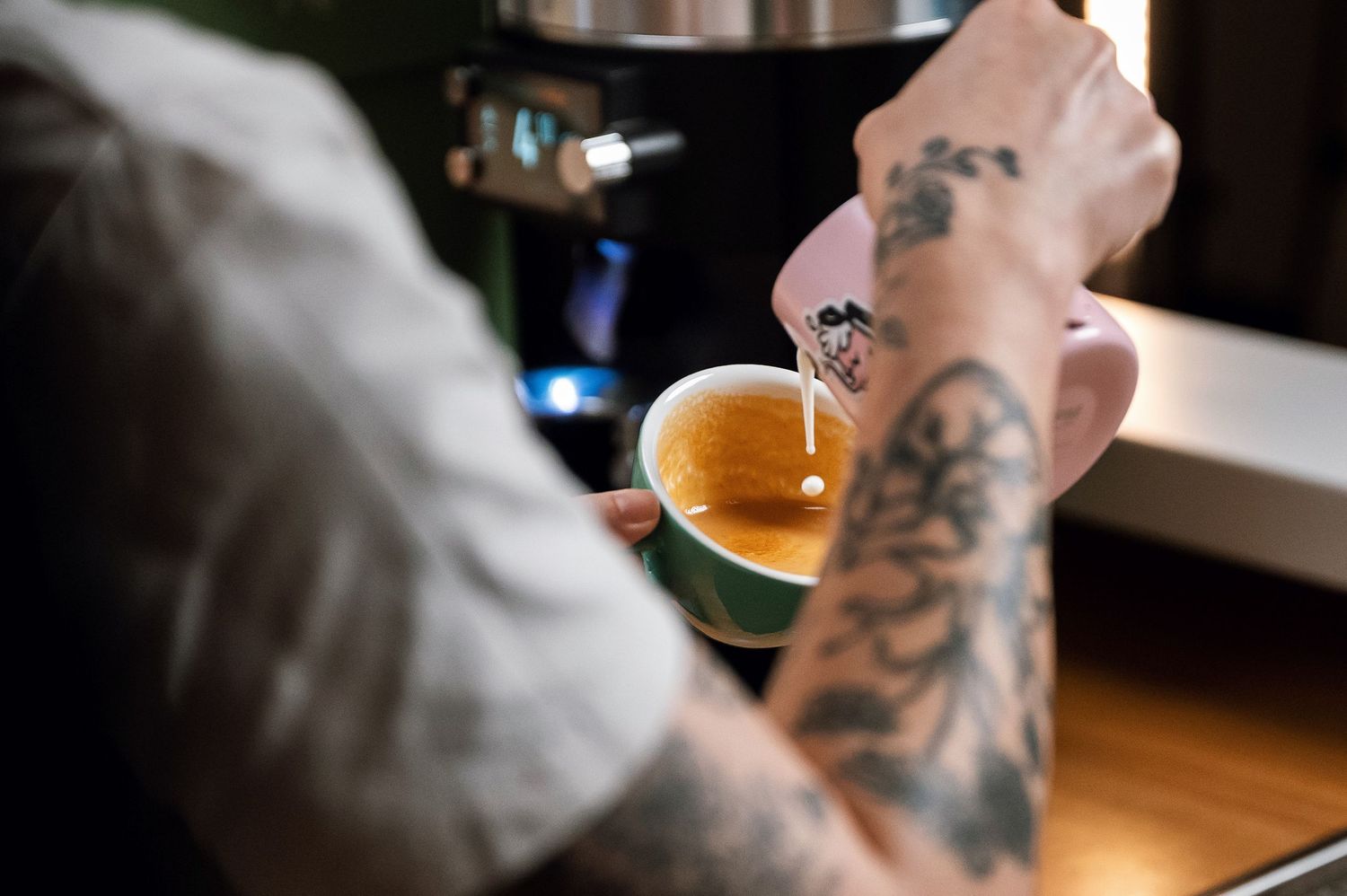 Tattooed barista pouring hot water into a green ceramic espresso cup at a coffee bar. Bailies Coffee Roasters