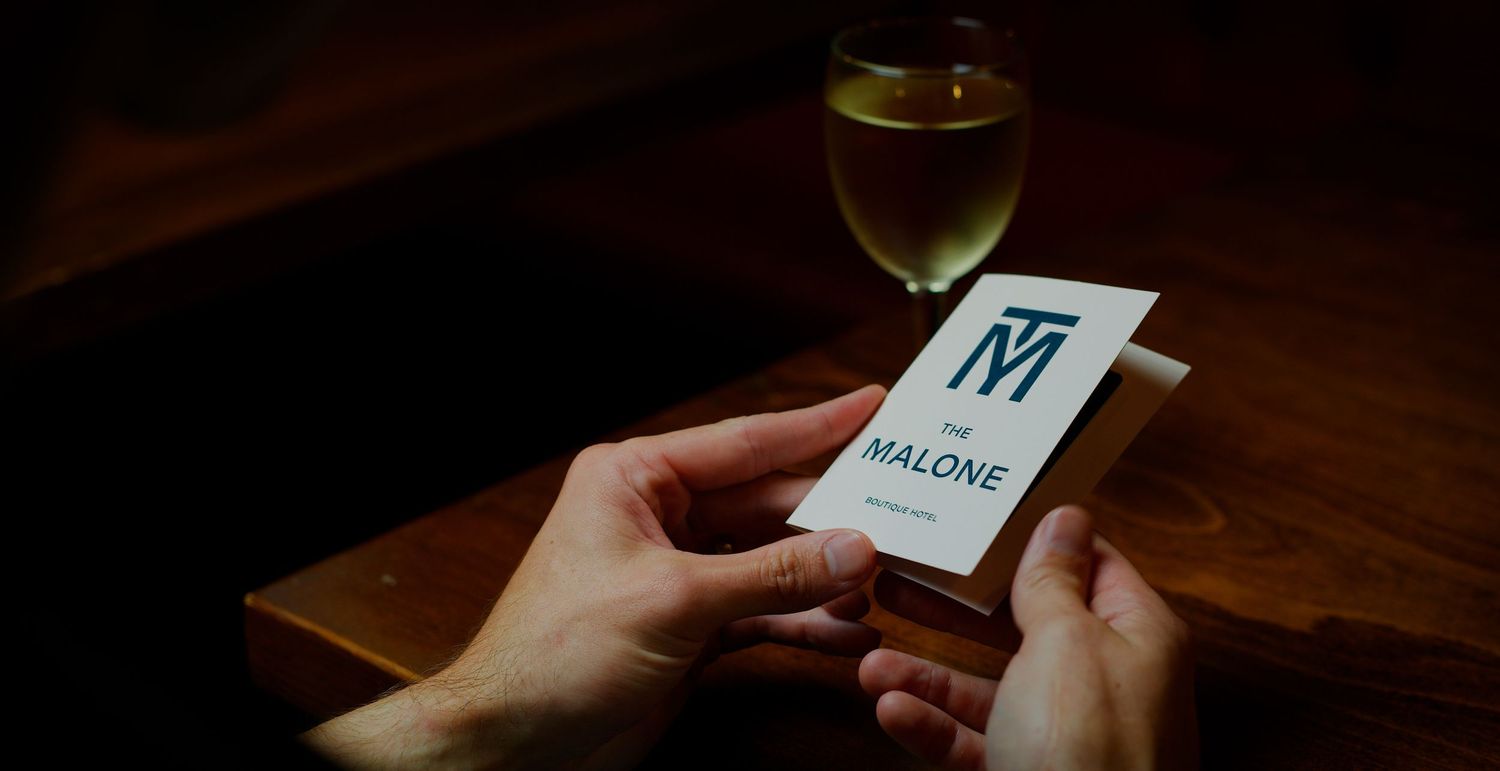 Hands holding a Malone branded business card near a glass of white wine on a wooden table. Kaizen Brand & Print