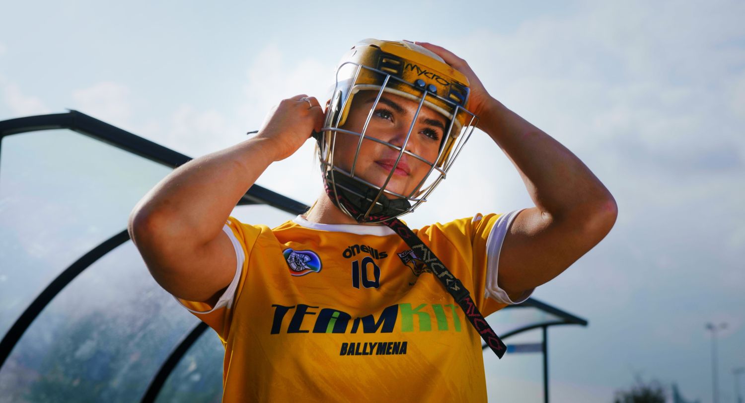 Female GAA player putting on a yellow helmet with Team Fit Ballymena jersey outdoors. Ulster GAA