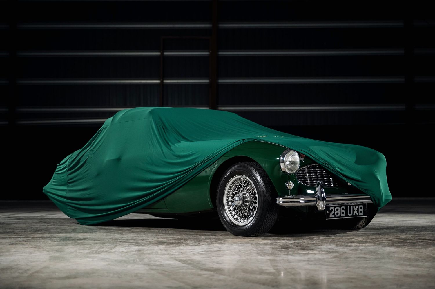 Vintage green sports car partially covered with a matching green car cover, showcasing wire wheels and classic front grille.