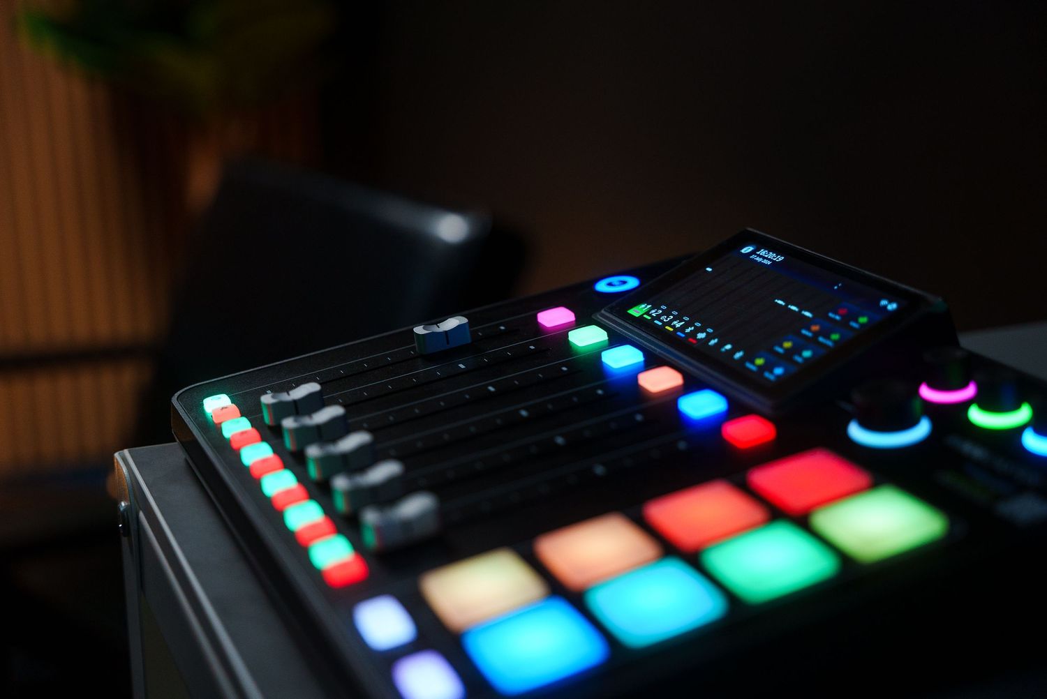 Colorful illuminated music production controller with RGB pads and faders in a dark studio.