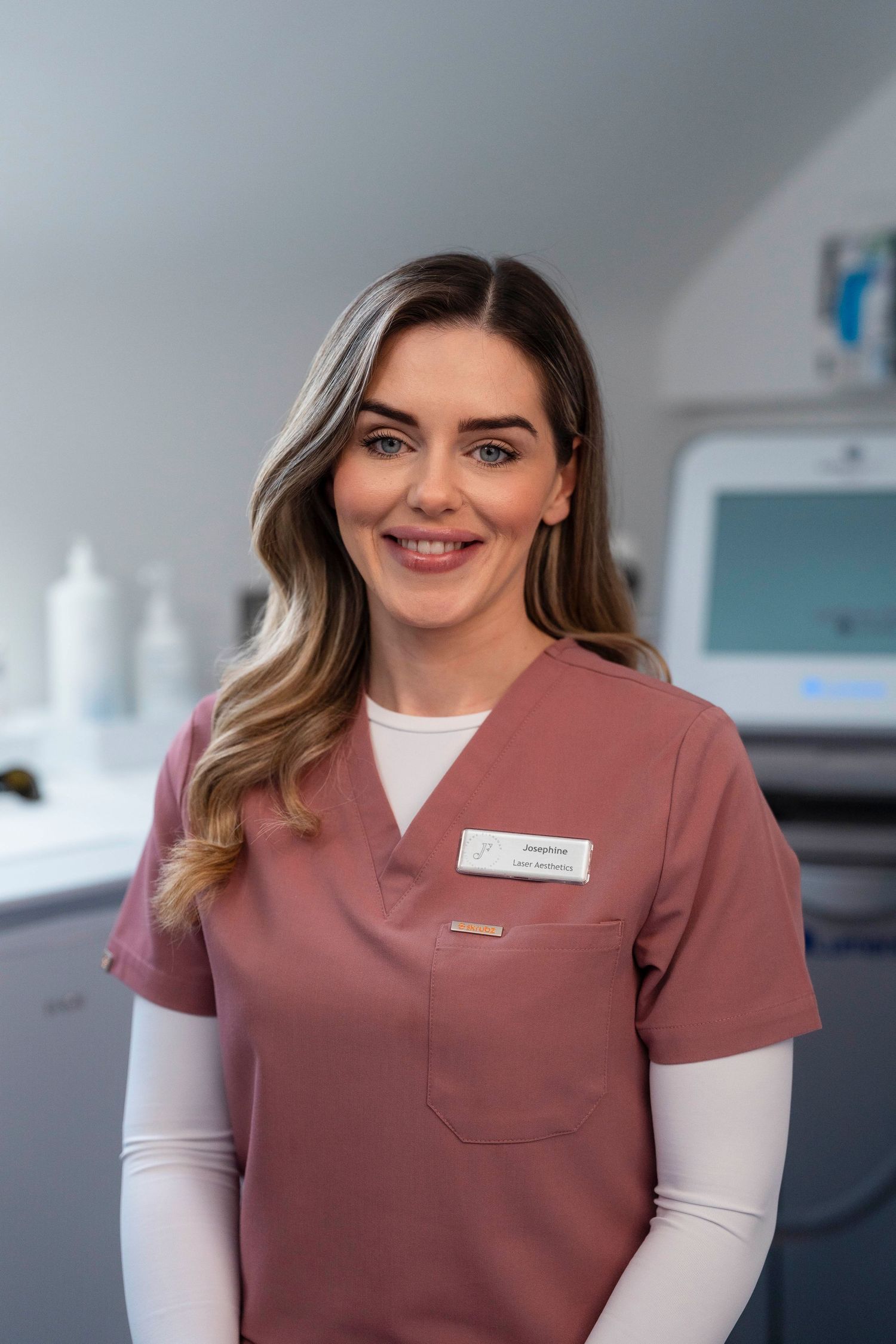 Smiling female beauty therapist in pink scrubs standing in a professional clinic room. Jenny Flanagan