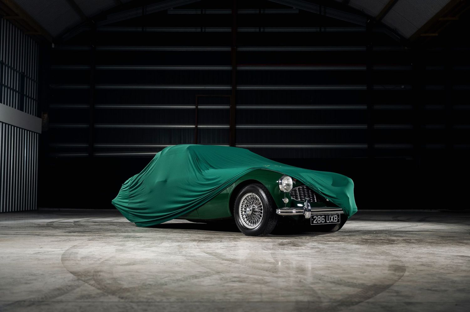 Classic vintage car covered with a green protective car cover stored in a dark garage. Motorist Collection