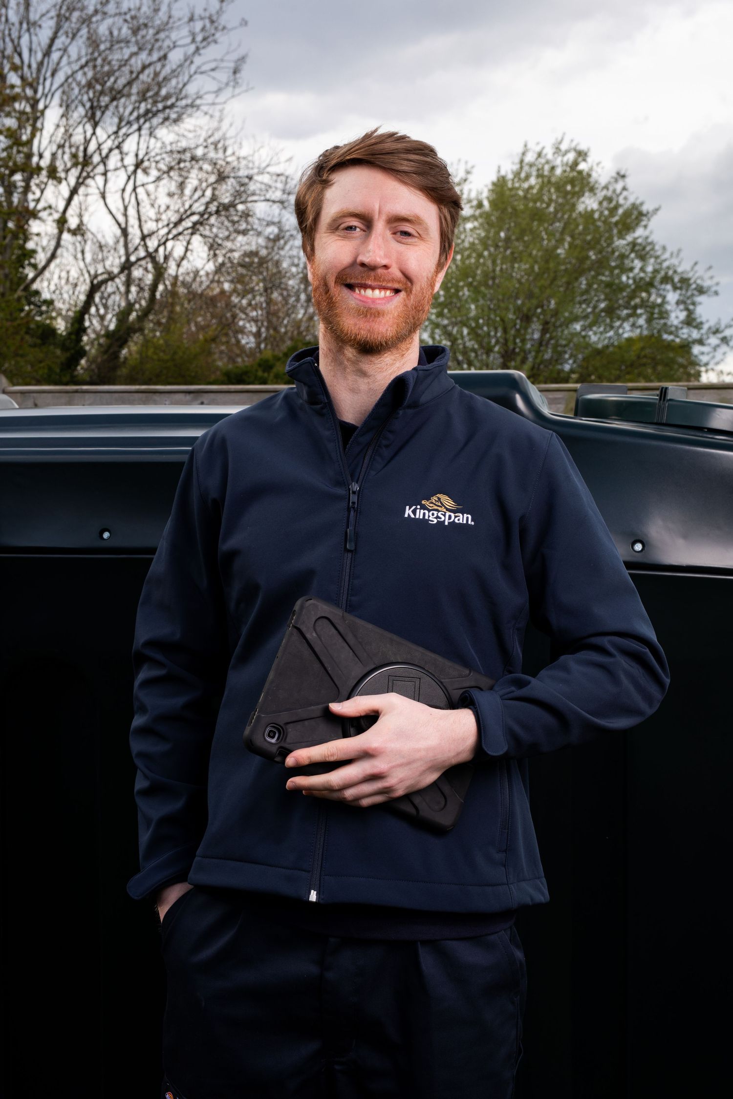 Smiling Kingspan engineer in navy uniform holding tablet standing outdoors beside dark storage tank. Kingspan Water & Energy