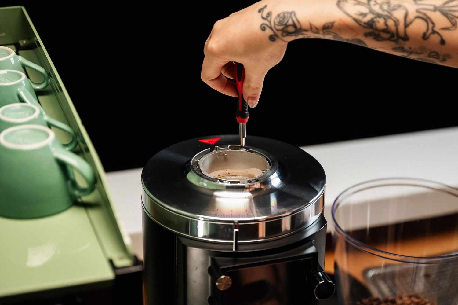 Tattooed hand adjusting a lid on a sleek chrome coffee grinder in a cafe setting. Bailies Coffee Roasters