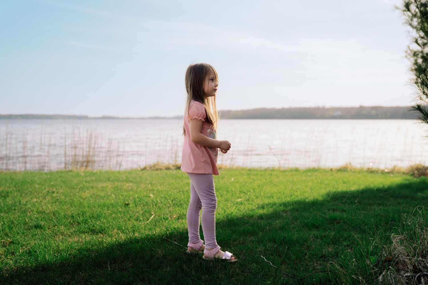 Young girl standing on green grass by a calm lake shore on a sunny day, wearing pink outfit.