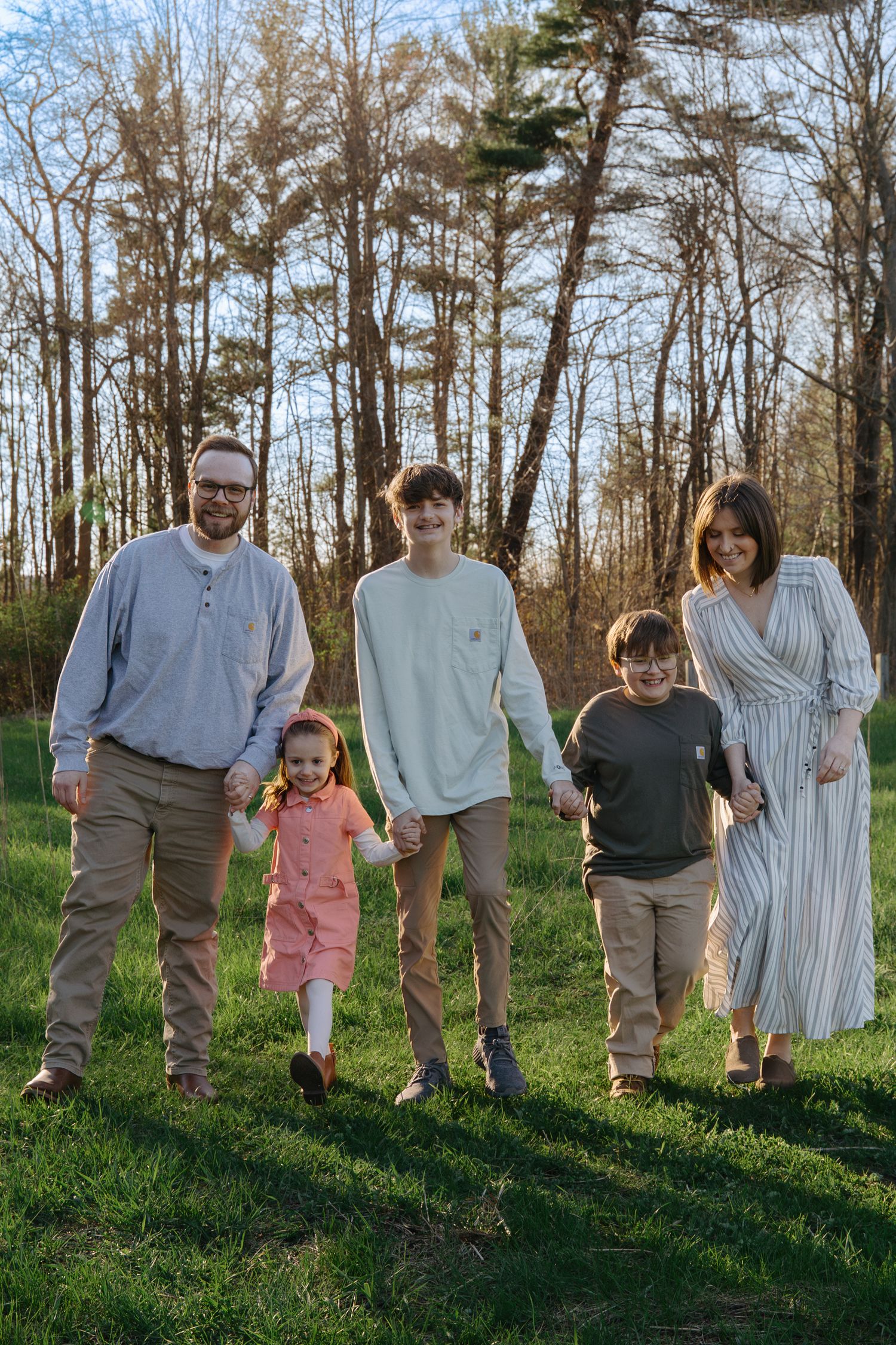 A happy family of five walks hand-in-hand on a grassy lawn with tall trees in the background during golden hour.
