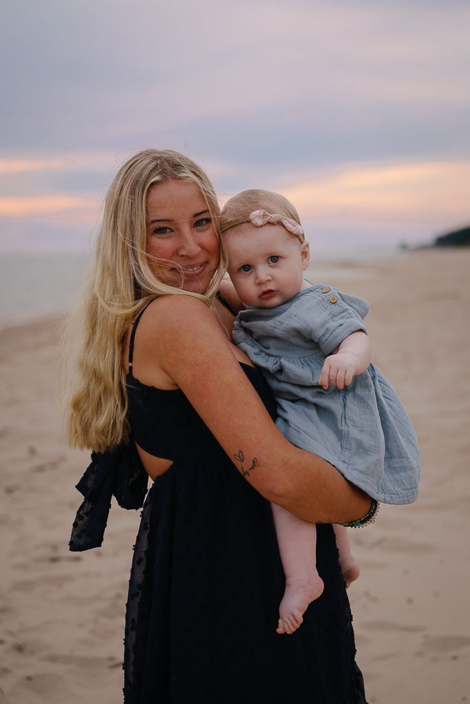 Mother and baby share tender moments on a beach at sunset with soft pink sky in the background.