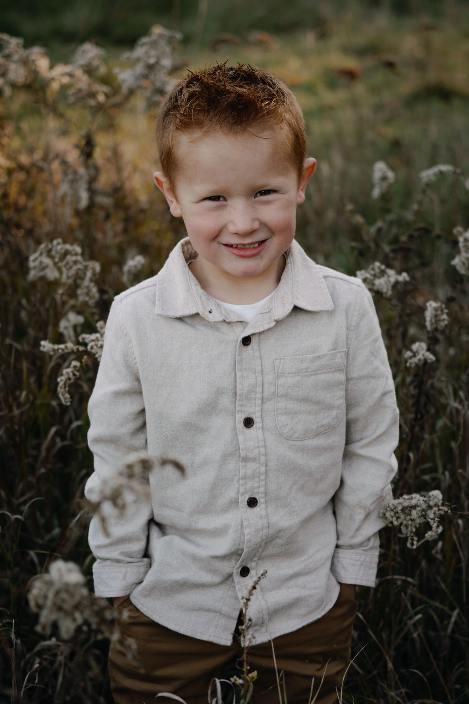 A child in a light button-down shirt smiles while standing among dried wildflowers at sunset.