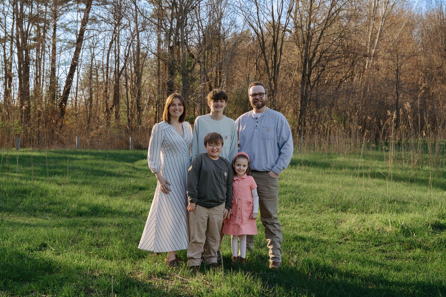 Family of five posing outdoors on green grass with trees in background during golden hour.