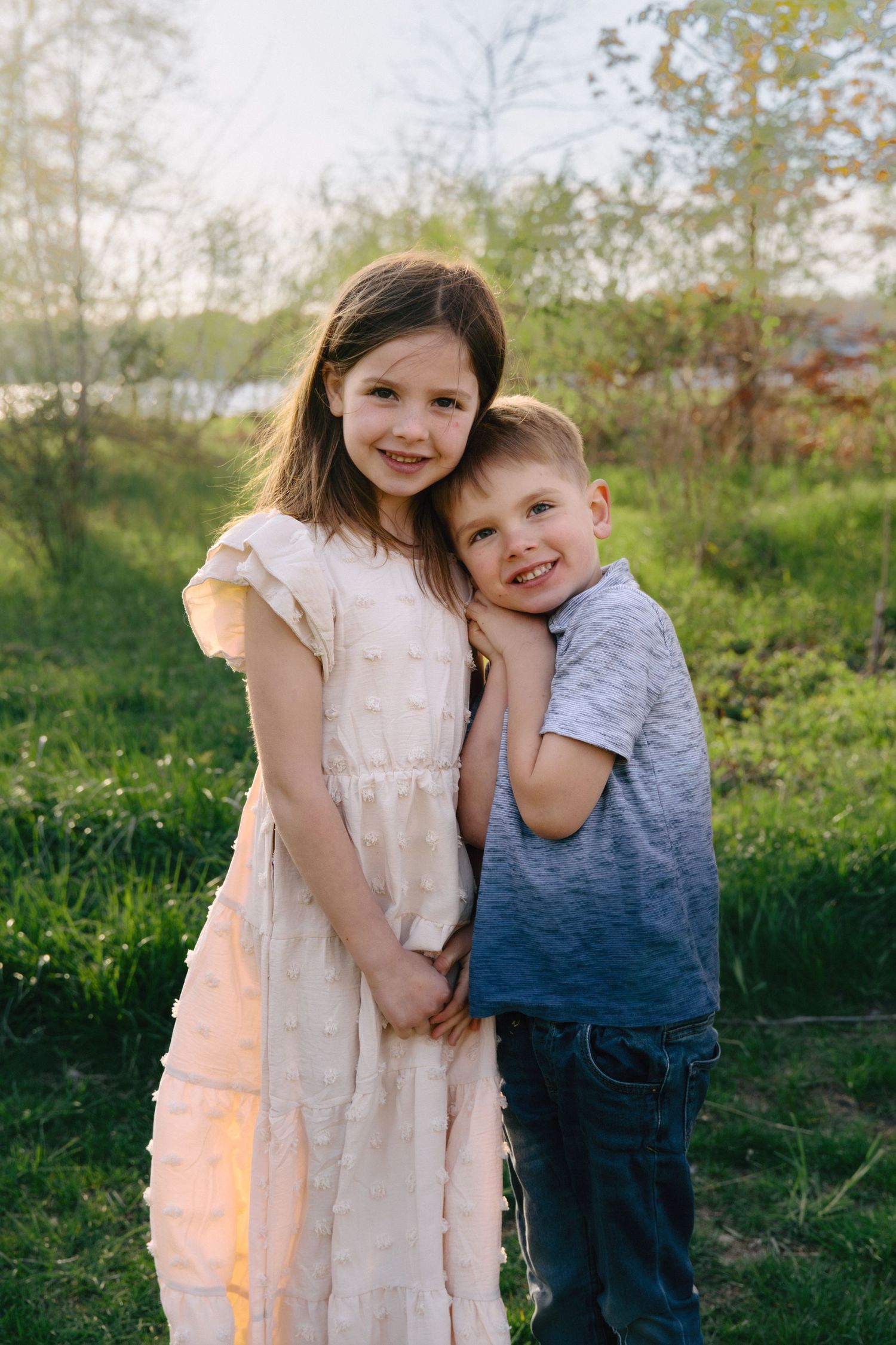 Two young siblings hugging outdoors in a sunny green park, girl in white dress and boy in gray t-shirt smiling.